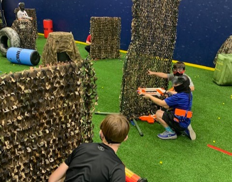 Kids in eye protection taking cover behind camouflage bunkers during a Nerf battle on the turf