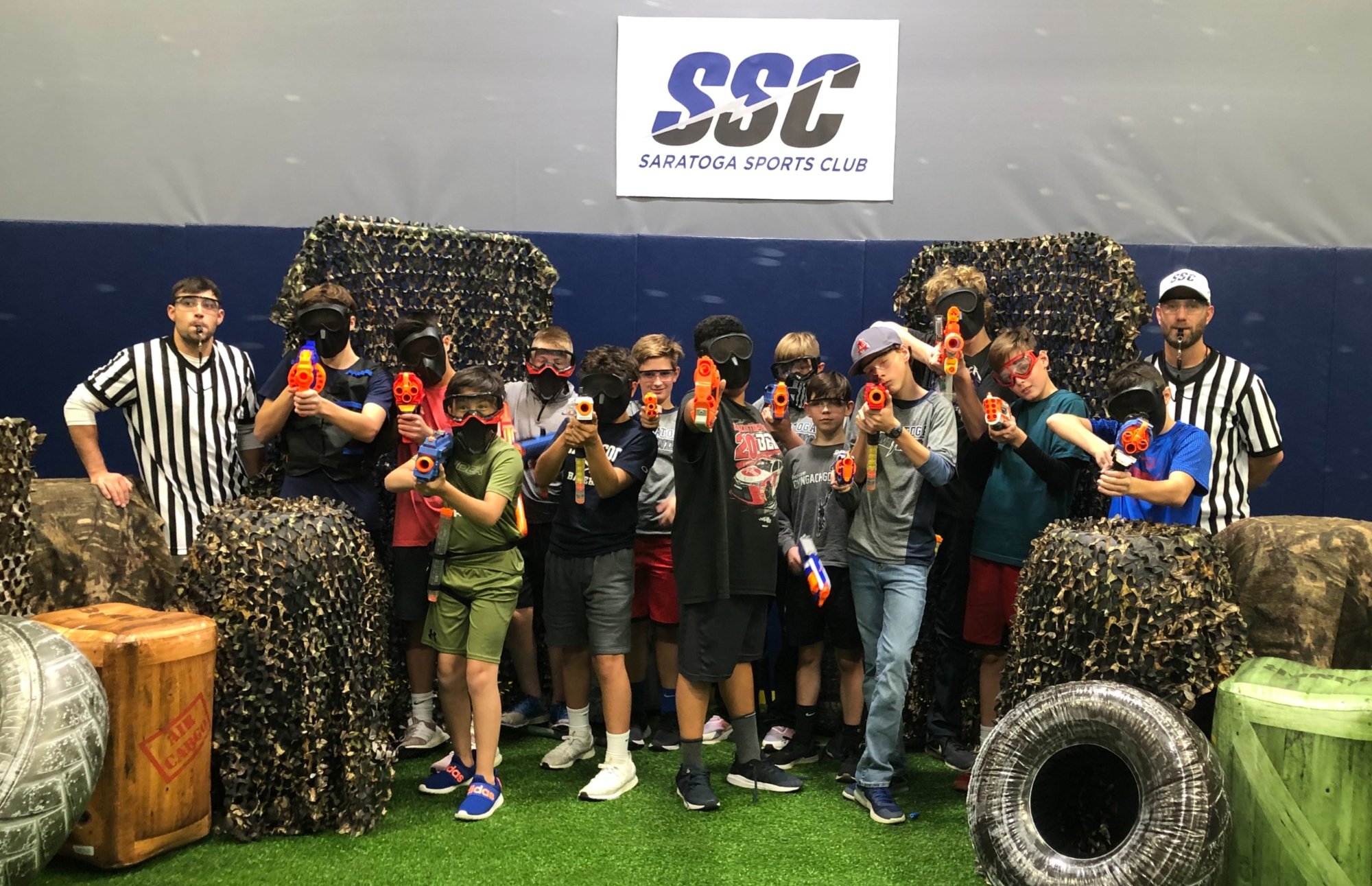 Kids with Nerf blasters and two referees in striped shirts, gathered under the Saratoga Sports Club sign on the turf arena