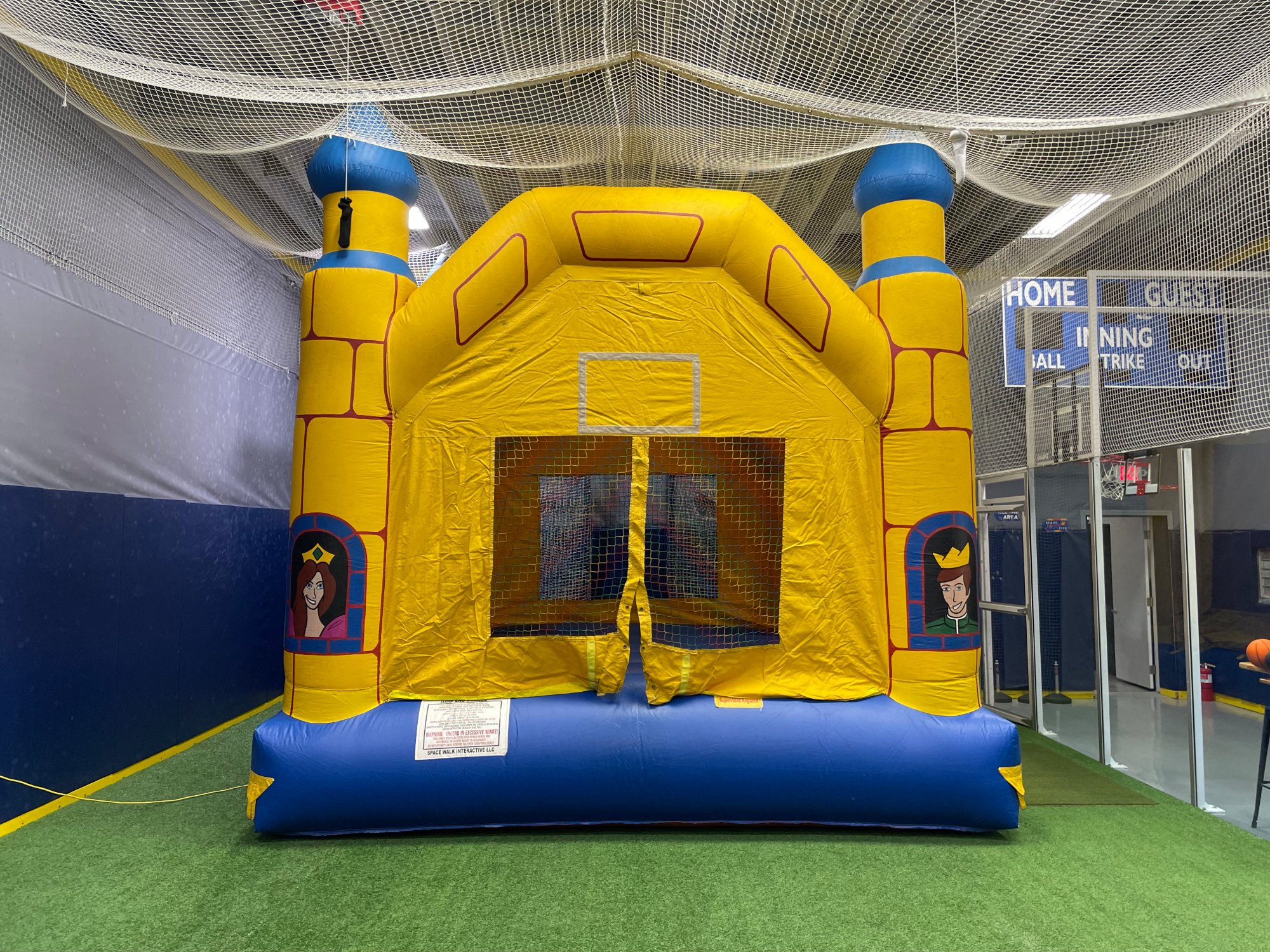 A yellow and blue bounce house set up on the turf at Saratoga Sports Club, next to the batting cage nets